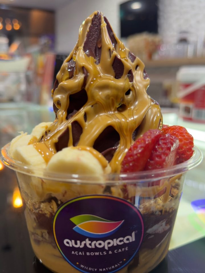 An açaí bowl with caramel sauce, banana and strawberry in a clear cup with the purple Austropical badge.