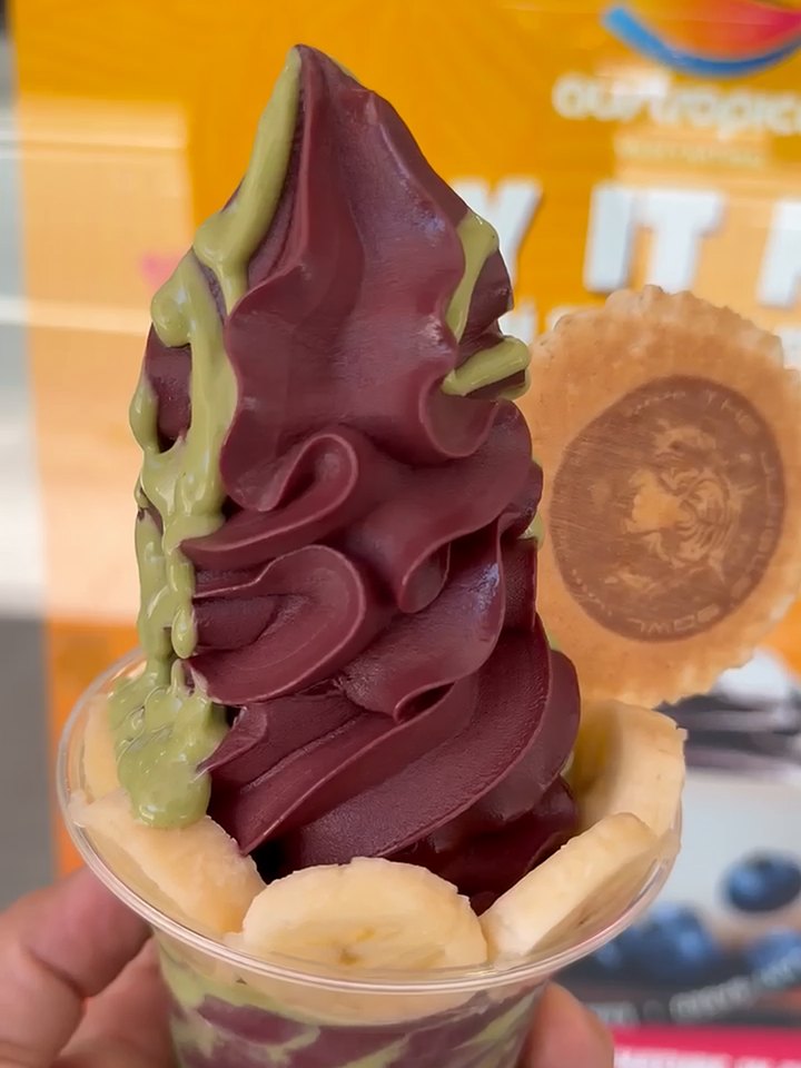 A tall swirl of açaí soft serve with pistachio sauce down one side, banana round the rim and a wafer, held up in daylight at the Austropical café in Sydney.