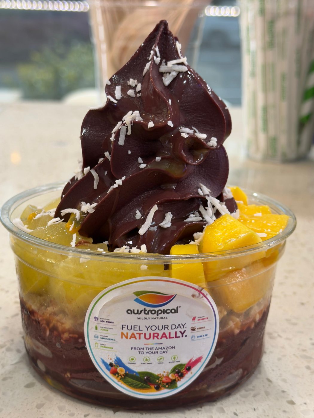 A swirl of dark açaí with shredded coconut, ringed with mango cubes and pale yellow fruit, on the café counter.