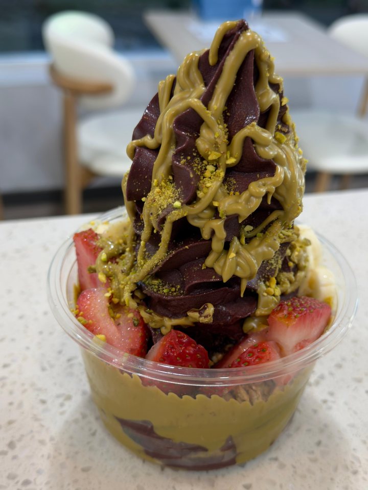 A tall swirl of açaí soft serve under pistachio sauce with strawberries, by the window at the Austropical café in Sydney.