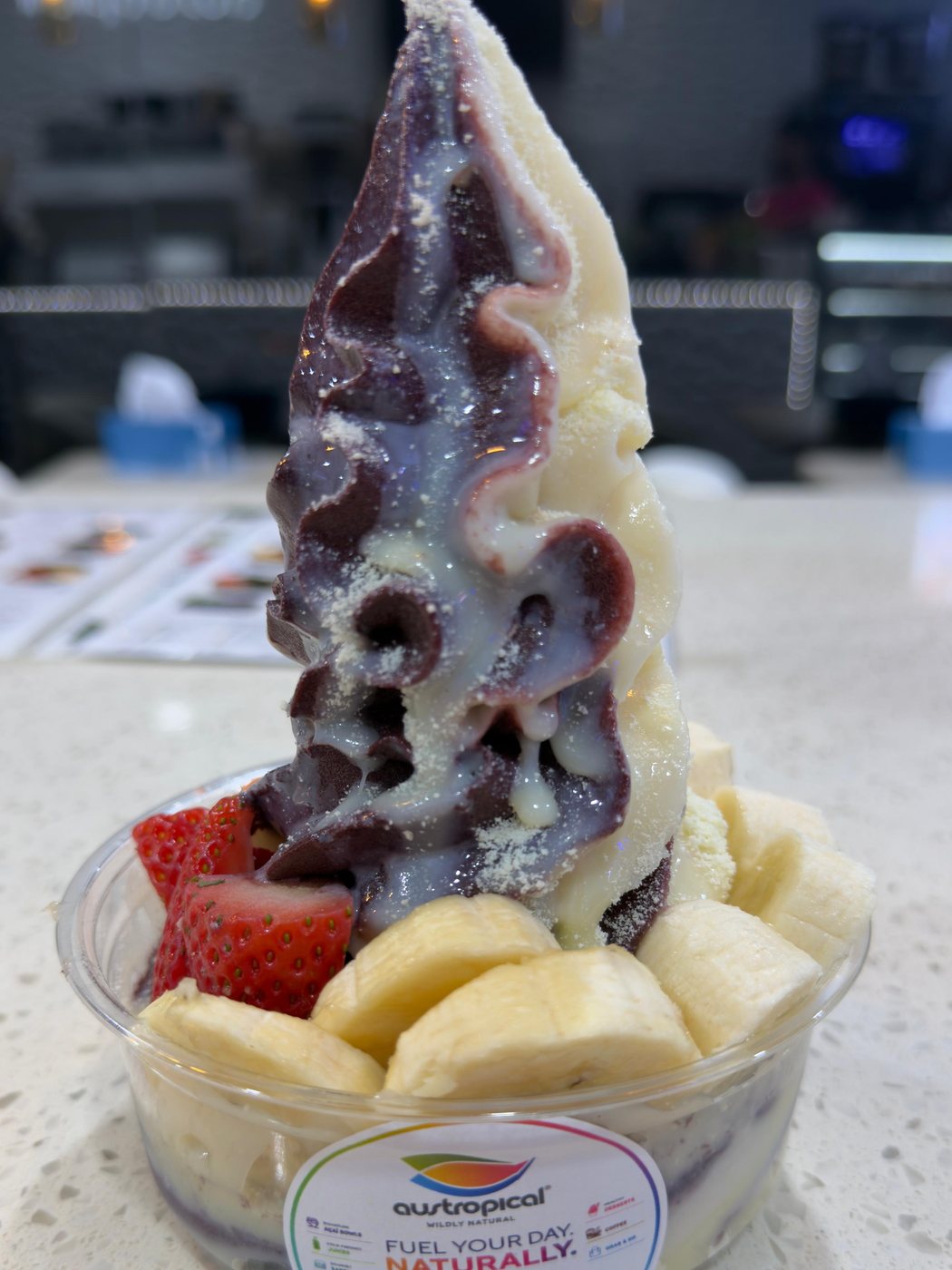 A tall two-tone swirl of açaí and a white soft serve under a white drizzle and a fine white dusting, over strawberries and banana, in a branded cup on the café counter.