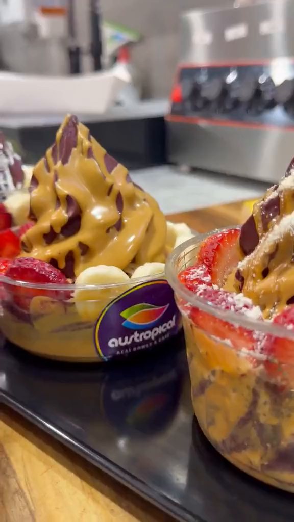 Finished açaí bowls on a black tray at the counter: caramel, chocolate and pistachio tops over banana and strawberry.