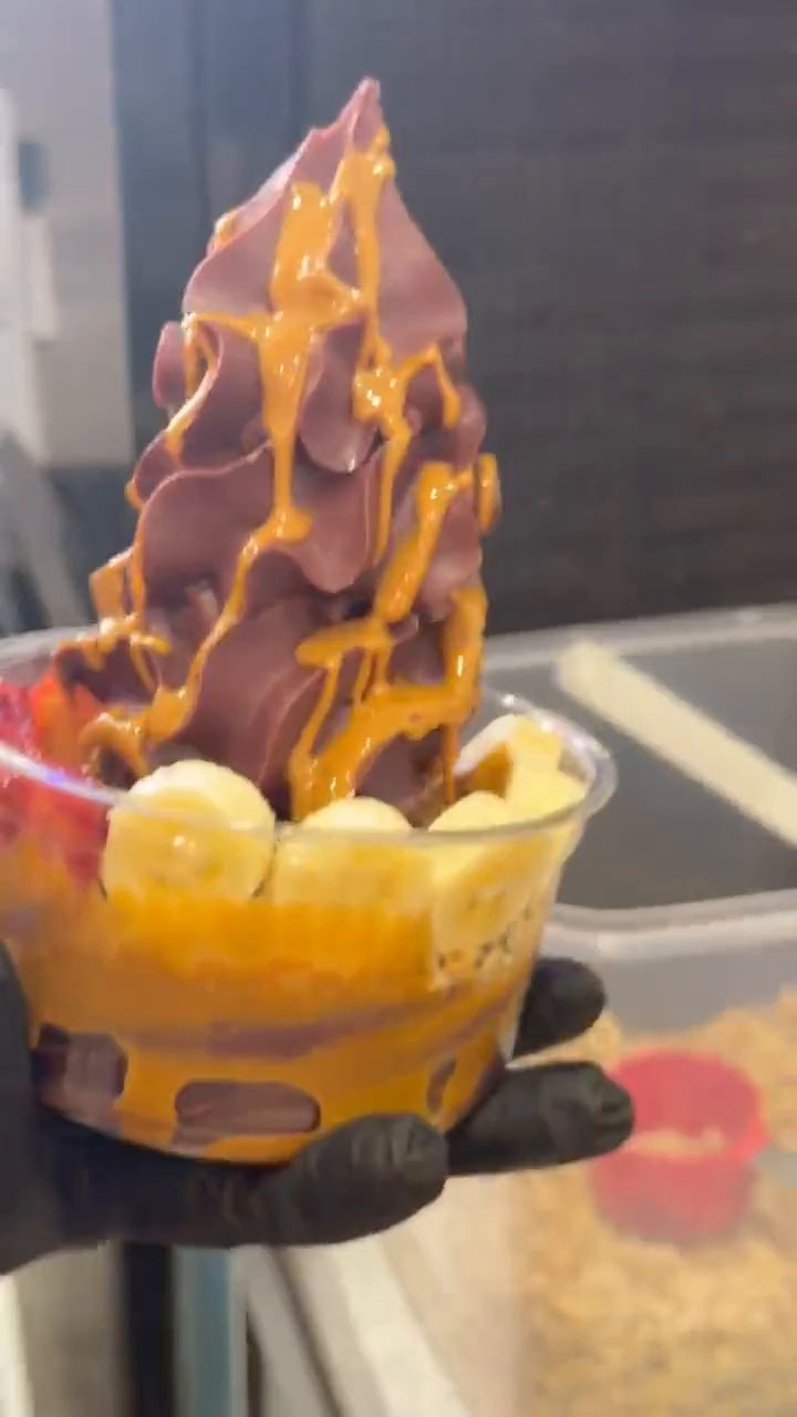 Caramel sauce poured over a tall swirl of açaí in a clear cup, held over the counter.