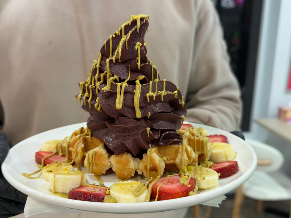 A waffle under a tall swirl of açaí soft serve with pistachio sauce, banana and strawberries, carried out on a plate.