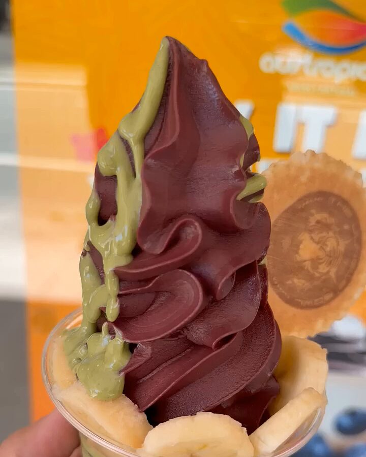 A hand holds a tall açaí soft-serve swirl with pistachio sauce, a banana collar and a round wafer, in front of an orange café poster.