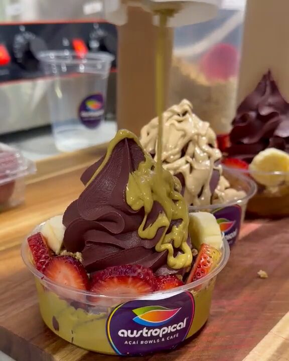 Pistachio sauce poured from a squeeze bottle over an açaí soft-serve bowl with strawberries and banana, in a cup with the purple Austropical badge.