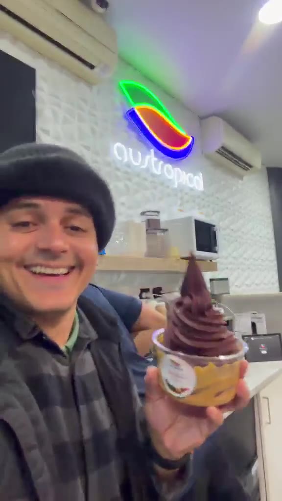A smiling man holds a tall açaí bowl at the counter beside a staff member and the neon Austropical sign.