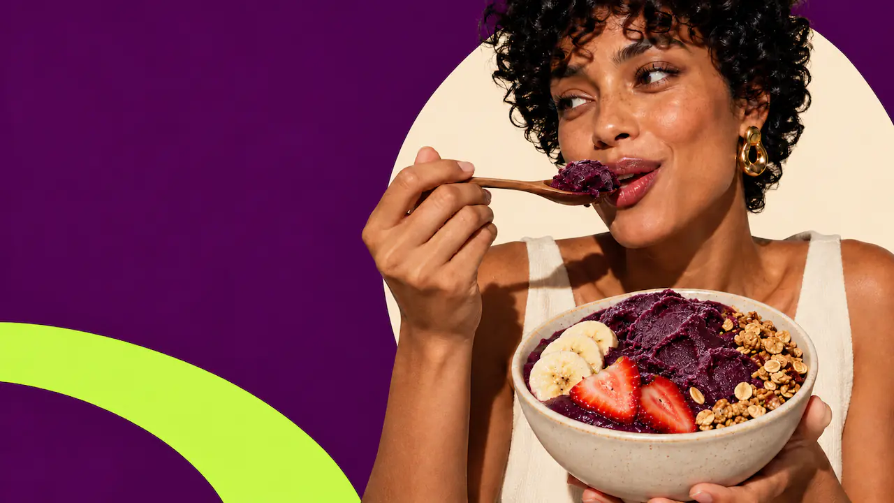 A woman with curly hair smiles as she takes a spoonful of açaí from a bowl topped with banana, strawberry and granola.